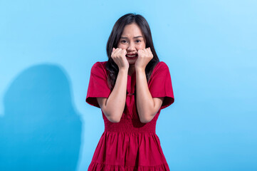 An Asian woman wearing a red dress, expressing fear with her hands clenched near her face and wide eyes, showing a frightened or anxious reaction. She stands against a light blue background