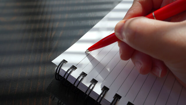 A man writes with a red pen in a notebook on a spiral, the sun's rays fall on the paper. The hand holds a pen and is ready to take notes, creating a feeling of work or study. A blank sheet of paper gi