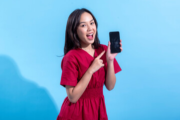 An Asian woman wearing a red dress is smiling and pointing at a black smartphone she is holding in her right hand. The light blue background complements her vibrant red outfit