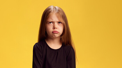 Photo of little girl with long blonde hair crosses her arms while pouting in frustration against yellow studio background. Concept of bad mood, childhood, kid's emotions. Ad
