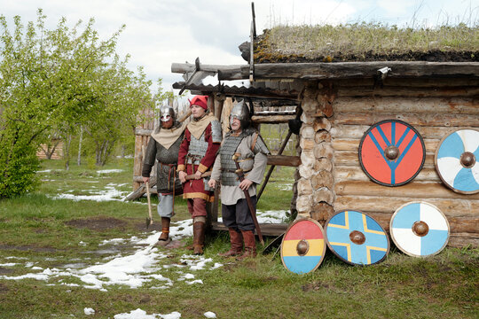 Group of historical reenactors in medieval warrior costumes standing in front of a wooden hut with shields on display around. Sparse snow patches on ground contrast with green surroundings