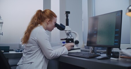 Fototapeta premium Medical science lab: Female microbiologist looks under microscope. Professional scientist does analysis of sample using high-tech equipment. PC screen shows image of cells or bacteria. Microbiology.