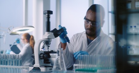 Scientists in white coats work in advanced laboratory. African American microbiologist examines...