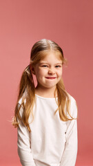 Smiling softly, girl bites her lip in shy but playful gesture. Sweet and innocent moment against pastel pink background. Concept of human expression, shyness, playful innocence, childhood moments.