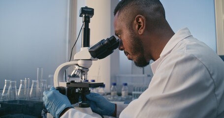 Medical science lab: Dolly shot of African American male microbiologist looking under microscope, analyzing test sample. Professional scientist, chemist uses high-tech equipment. Scientific research.