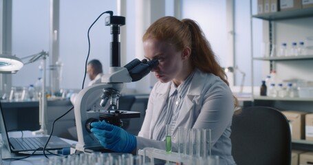 Female scientist or chemist wearing protective glasses to analyze and study new sample under microscope, uses laptop computer. In the background doing analysis. Modern medical science laboratory.