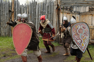 Group of medieval reenactors in period costumes engaging in mock battle holding shields and weapons in hand-made wooden fort setting enjoying historical play