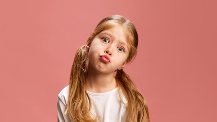 Cute little girl creates pouty, playful expression, lips puckered and her head tilted, giving off sense of silly defiance against pastel pink background. Concept of human emotions, childhood, positive