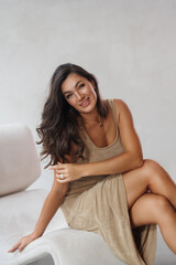 Obraz premium A beautiful tall brunette in a gold knitted dress. Spectacular volumetric styling. Portrait of a smile. The woman sit on beige sofa near window