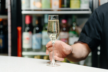 Cropped photo of young bartender taking in hand glass of champagne behind bar counter in bar, closeup view, alcohol bottles in background, nightlife concept