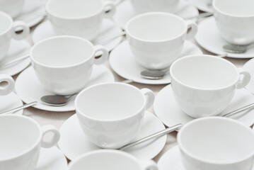 white empty porcelain cups on saucers with spoons in several rows on white tablecloth, serving tables for banquet in restaurant or cafe, pattern, background
