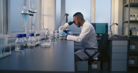 Medical science lab: African American male microbiologist looks under microscope, analyzes test sample. Brilliant scientist uses high-tech equipment. Female lab worker takes test tubes from the table.