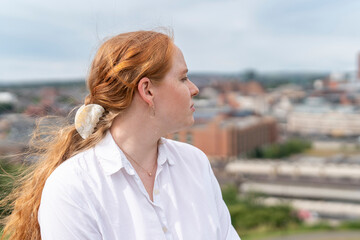 A young woman with long red hair gazes thoughtfully over the city skyline on a cloudy afternoon