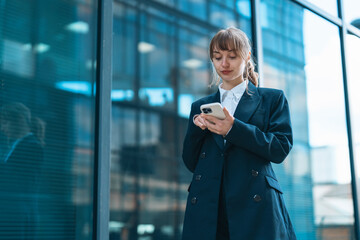 A young professional engaging with her smartphone outside a modern office building in bright daylight