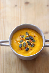 Bowl of creamy pumpkin and potato soup, with pumpkin seeds and chilli flakes toppings. Selective focus, wooden background.