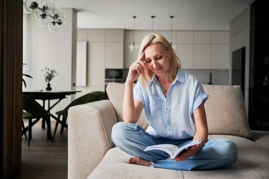 Woman sitting comfortably on couch, deeply engaged in reading book. Cozy living space with modern aesthetic on background. Person enjoying quiet moment of solitude, immersed in reading.