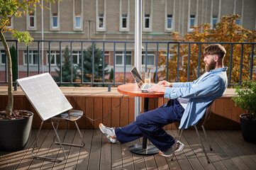 Man works on laptop at outdoor round table, with glass of iced coffee. Solar panel nearby,...