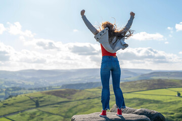 Joyful young woman celebrates on a rocky outcrop, overlooking a breathtaking green valley at midday