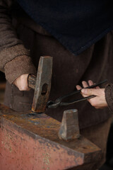 Close-up view of person's hands hammering metal on an anvil, illustrating traditional blacksmithing techniques. Rustic setting adds authenticity to craft