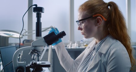Female scientist or chemist wearing protective glasses to analyze and study new sample under microscope, uses laptop computer. In the background doing analysis. Modern medical science laboratory.
