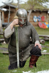 Medieval warrior dressed in chainmail and helmet kneeling on grass holding large sword in an...