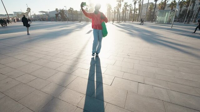 Young African American woman using smartphone while walk on Barcelona seashore, staying connected on the go. Cheerful black female tourist in Casual attire with headphones using 5G roaming connection
