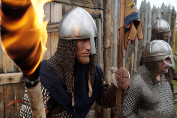 Group of reenactors wearing medieval armor standing in front of wooden fort holding torches while...
