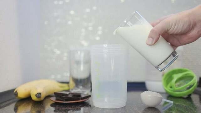A male hand pours milk and adds whey protein into a shaker, in the process of making a protein cocktail or smoothie drink in a kitchen, captured in slow motion