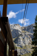 A scenic view of a cable car ascending a majestic mountain, framed by a balcony overlooking the valley. The vibrant colors of the car contrast with the rugged cliffs and lush greenery below.