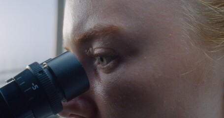 Close up of eyes of female doctor looking under microscope while conducting pharmacological test on...