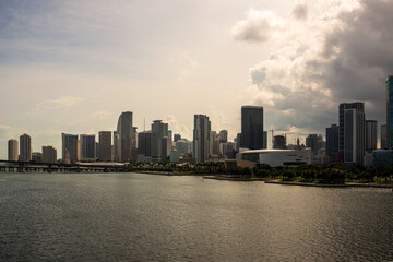 Fototapeta premium A modern city skyline with skyscrapers overlooking calm waterfront under a bright yet overcast sky. The contrast between the towering buildings and the water creates a dynamic urban landscape.