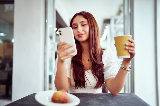 portrait young latina woman sitting in a cafe holding coffee cup and using smartphone	