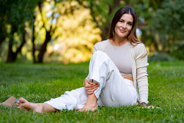 Beautiful woman is smiling while sitting barefoot on the grass in a park, enjoying a sunny day
