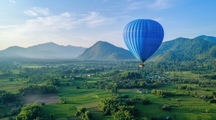 Fototapeta premium A bright blue hot air balloon floating above a green valley, with a clear sky and mountains in the background.