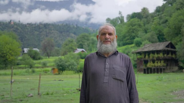 A mid shot of old Indian Kashmiri man or male standing outdoors in traditional attire in the green meadow looking at the camera smiling with scenic green mountain valley and clouds in the background