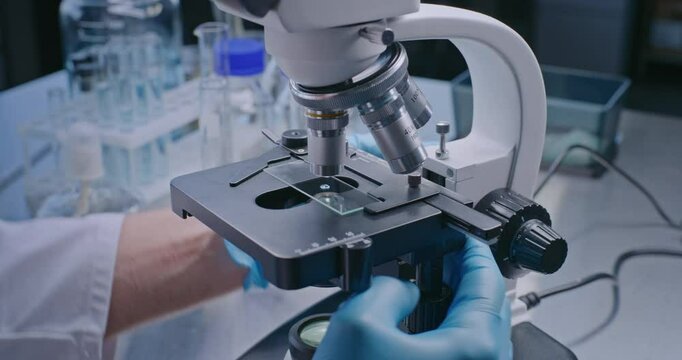 Medical science lab: Close up of professional microbiologist or scientist in gloves putting slide with test sample under microscope for analysis, setting up device. High-tech laboratory equipment.