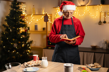 elderly man with cookbook in kitchen decorated for Christmas preparing to bake gingerbread cookies at home on Christmas Eve or New Year.