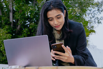 Fototapeta premium Businesswoman using mobile smart phone during working on laptop computer outdoors. Asian woman using smartphone searching the information with laptop, internet technology concept.