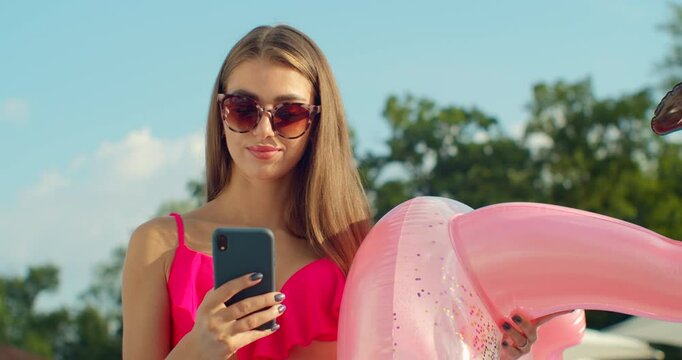 Summer girl using smartphone and sunbathing at swimming pool. Female model relaxing outdoors at sunny day.