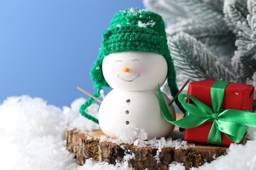 Cute decorative snowman, gift box and fir tree branches on artificial snow against light blue background, closeup