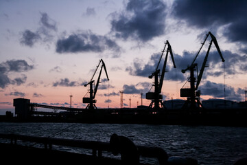 Fototapeta premium Three industry crane silhouette at a seaside port with dark clouds and pink sunset gradient