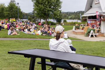 Mother is holding her baby while enjoying an outdoor show in a park, sitting on a picnic table....