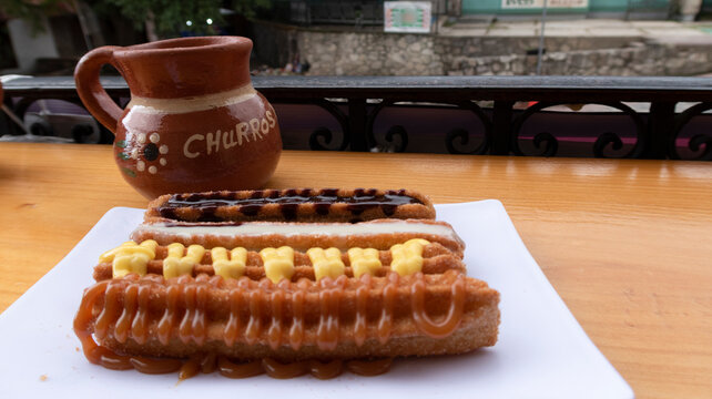 tres churros de frente con lechera, chocolate y cajeta en plato con caf&eacute; en jarro al fondo