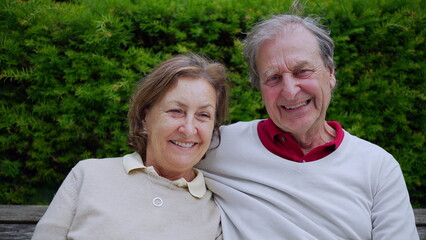 Elderly couple sitting closely together on a bench, smiling and enjoying a joyful moment outdoors, surrounded by lush greenery, capturing the warmth of their relationship