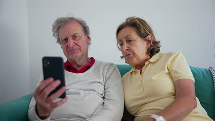 Elderly couple sitting together on a sofa, focused on a smartphone screen, sharing a moment of connection as they engage with technology in the comfort of their home