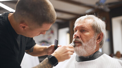 Senior man visiting barber shop. Male barber with tattooed arm carefully trimming beard, using electric clipper and comb. Concept of male beauty, barbershop, hairstyle, salon, hairdresser