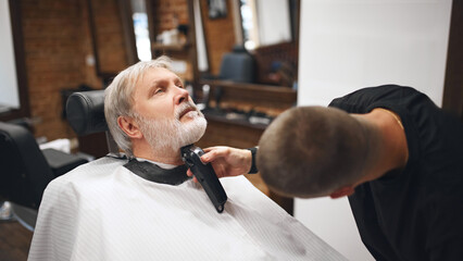 Barber using clipper to finish detailing client beard, focusing the contours of chin. Elderly man...
