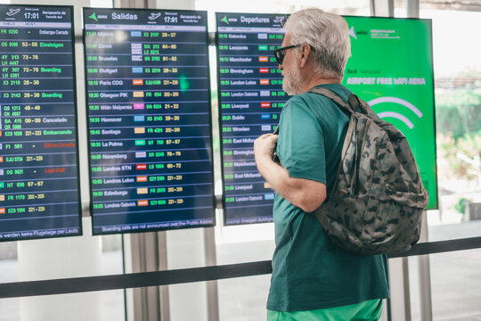 Senior bearded man travels at airport with backpack looking at time table display. Rear view of elderly male walking towards gate to board. Happy traveler enjoying trip and vacation