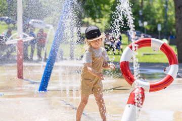 Young boy wearing a baseball cap is playing and splashing in the water jets of a colorful splash pad on a sunny summer day. Selective focus