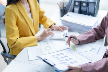 Two businesswomen in a modern office discussing financial data, using a calculator and charts. Working together, showcasing teamwork skills in a corporate meeting focused on growth and success